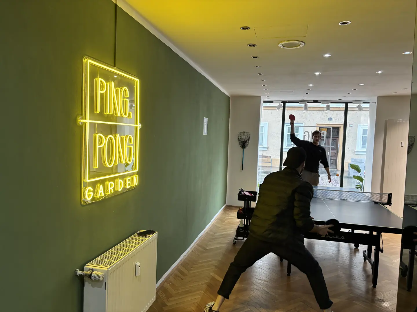 Neon-Schriftzug „Ping Pong Garden“ an der grünen Wand, dahinter ein Spieler beim Ballwechsel
